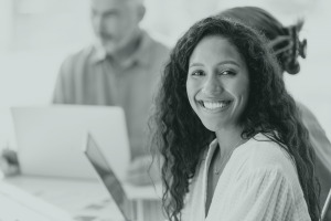 Femme souriant dans un bureau, avec deux personnes dans le fond devant leur ordinateur