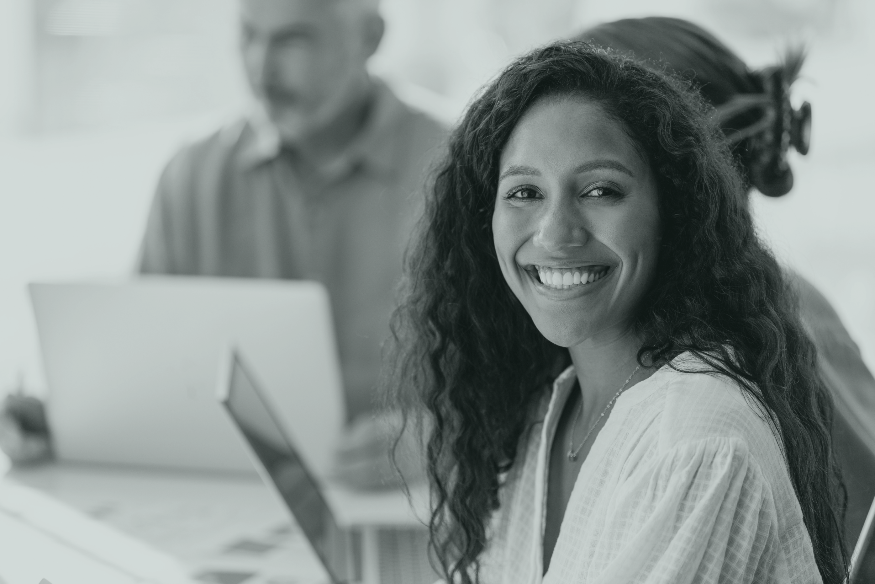 Woman smiling in an office, with two people in the background in front of their computers