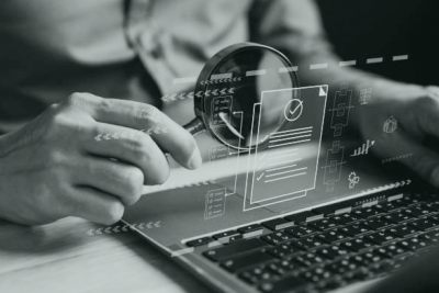 Muvraline_Bancassurance_Analyse_Texte Vue rapprochée d'une main tenant une loupe devant un ordinateur avec des illustrations de documents, rouages et courbes