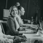 Three people in profile in front of their computers on a long desk, the one in the middle with her head turned and a big smile on her face